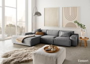 Moderne graue Rundecke mit Ottomane in einem hellen Wohnzimmer, seitliche Perspektive, dekoriert mit Kissen und Decke, umgeben von großen Fenstern und minimalistischer Kunst an der Wand.