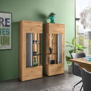 Zwei moderne Highboards aus Holz mit Glaseinsätzen, dekoriert mit Vasen und Geschirr, in einem stilvollen Wohnzimmer mit grüner Wand und großem Fenster, seitliche Perspektive.