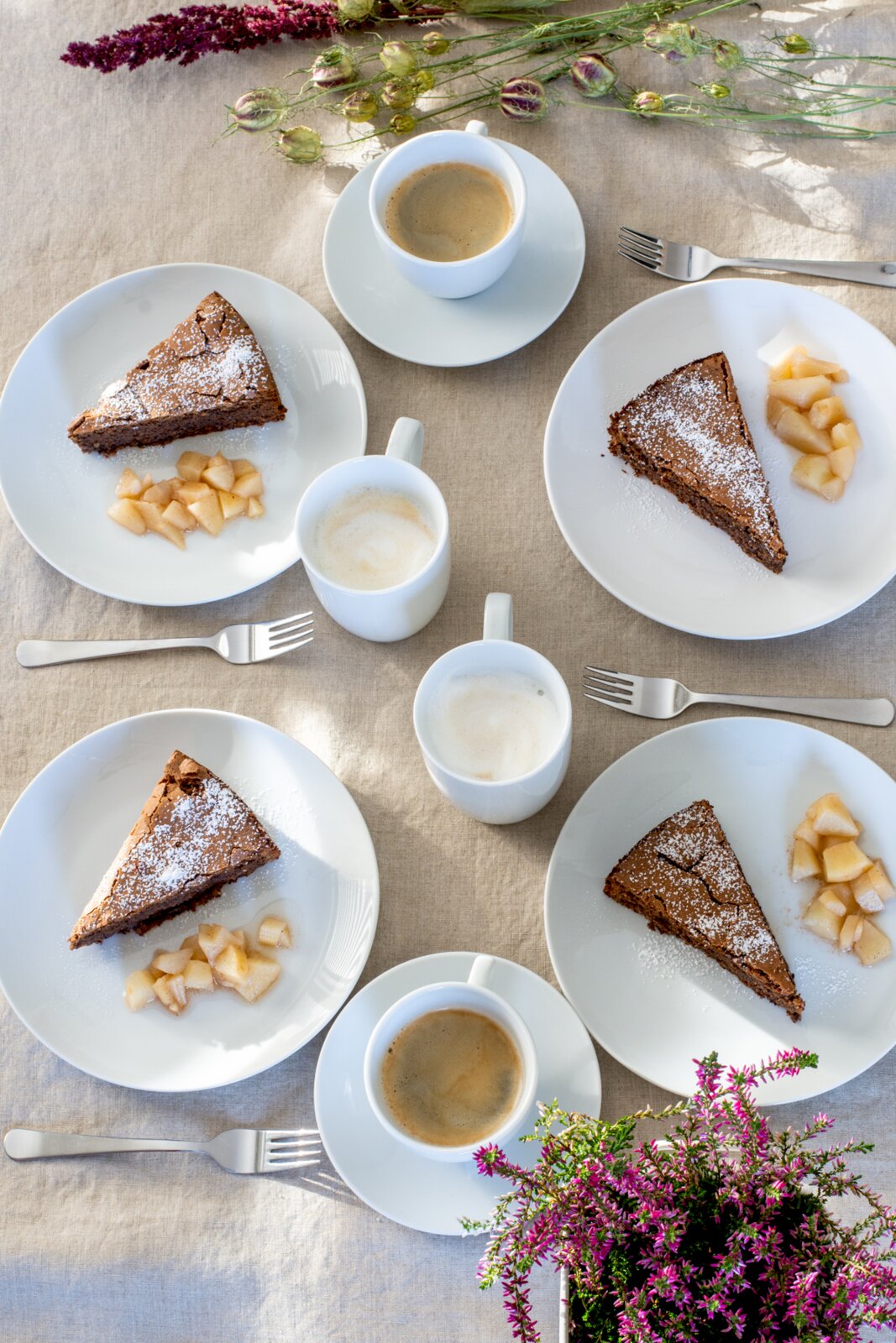 Draufsicht auf einen gedeckten Tisch mit vier Stücken Schokokuchen mit Puderzucker und Apfelstückchen auf weißen Tellern, dazu mehrere Tassen Kaffee und Gabeln auf hellem Leinentuch, dekoriert mit Blumen.