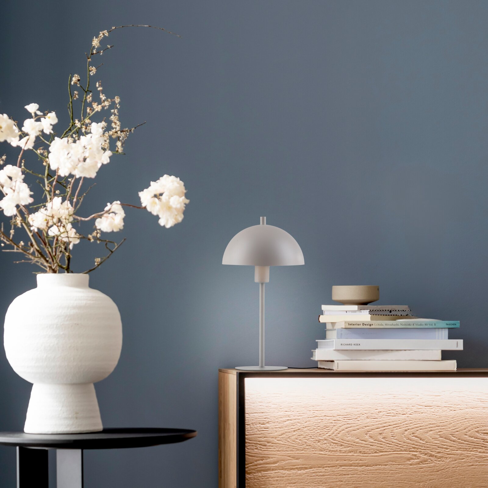 Minimalistische Tischleuchte in Weiß mit halbrundem Schirm auf Holz-Sideboard neben Bücherstapel und Keramikvase mit weißen Blüten, Frontansicht auf Augenhöhe