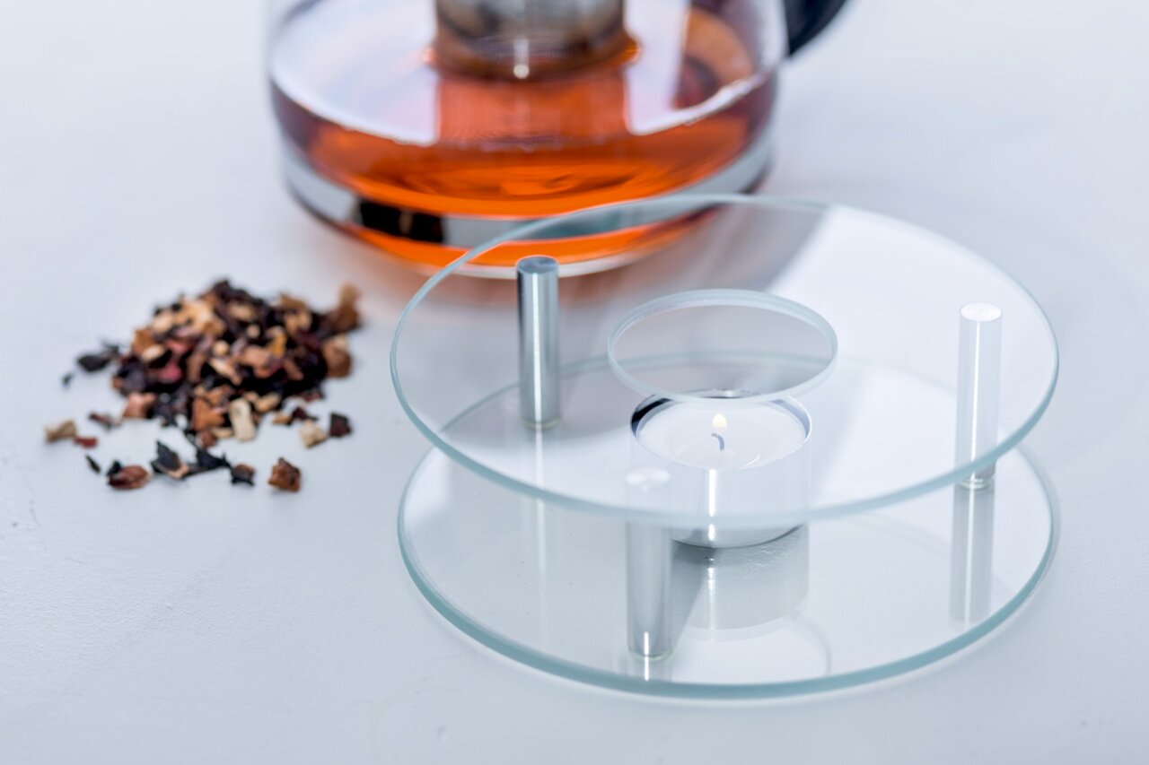 Glas-Stövchen mit Teelicht, seitliche Perspektive, neben einer Teekanne und losen Teeblättern.