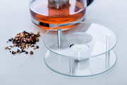 Glas-Stövchen mit Teelicht, seitliche Perspektive, neben einer Teekanne und losen Teeblättern.