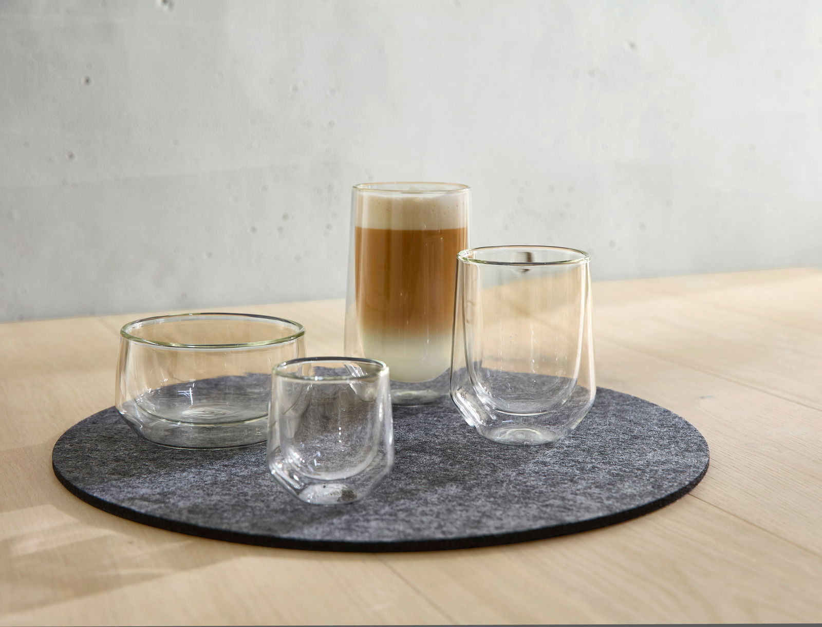 Vier doppelwandige Gläser auf einem runden Filzuntersetzer, perspektivisch von vorne rechts fotografiert, mit einem Latte Macchiato im größten Glas.
