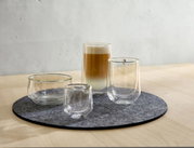Vier dubbelwandige glazen op een ronde vilten onderzetter, in perspectief gefotografeerd vanaf rechtsvoor, met een latte macchiato in het grootste glas.