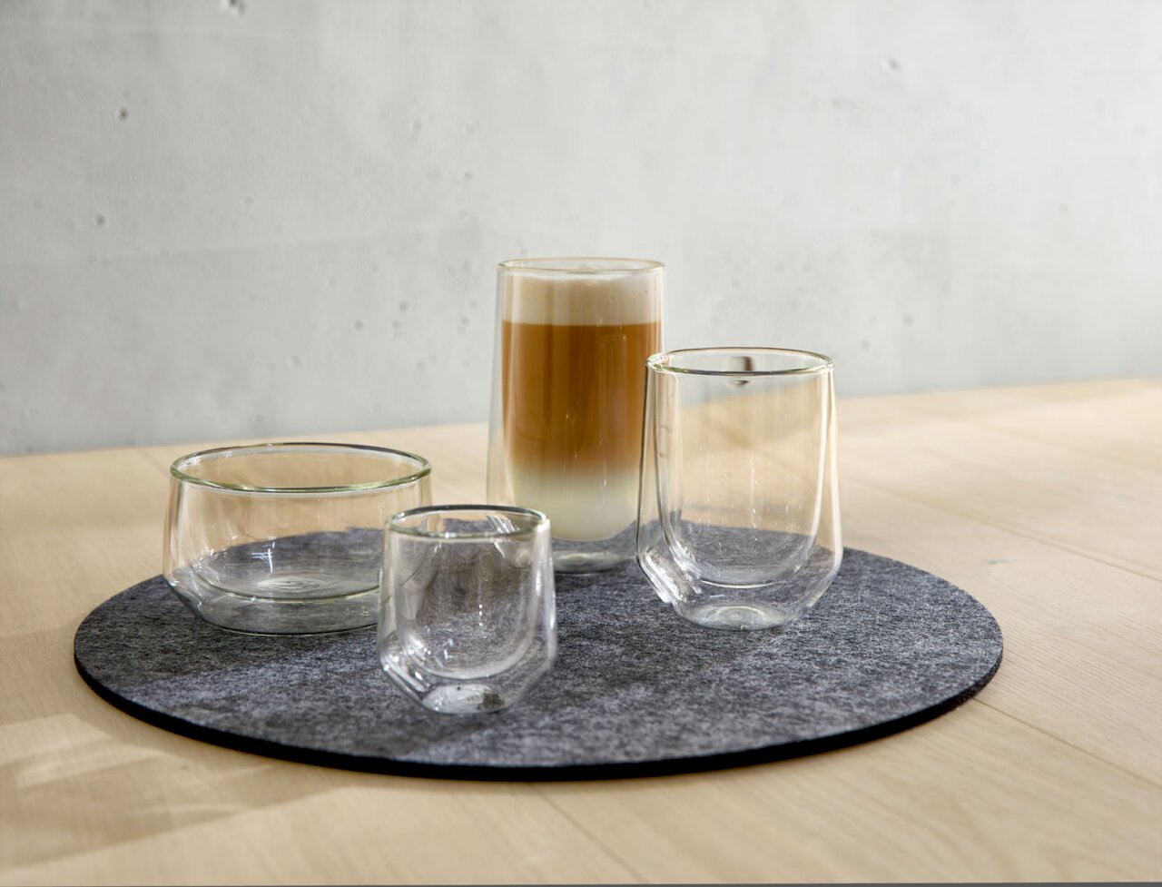 Vier dubbelwandige glazen op een ronde vilten onderzetter, in perspectief gefotografeerd vanaf rechtsvoor, met een latte macchiato in het grootste glas.