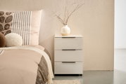 Weißer Nachttisch mit drei Schubladen und schwarzen Griffleisten neben einem Bett, darauf eine beige Keramikvase mit Zweigen; frontale Perspektive auf das Möbelstück.