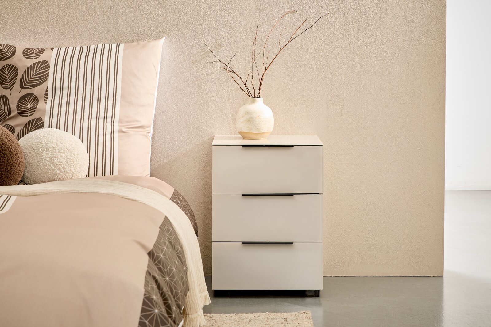 Novel Nachtkommode MALIN 2.0 Weißer Nachttisch mit drei Schubladen und schwarzen Griffleisten neben einem Bett, darauf eine beige Keramikvase mit Zweigen; frontale Perspektive auf das Möbelstück.