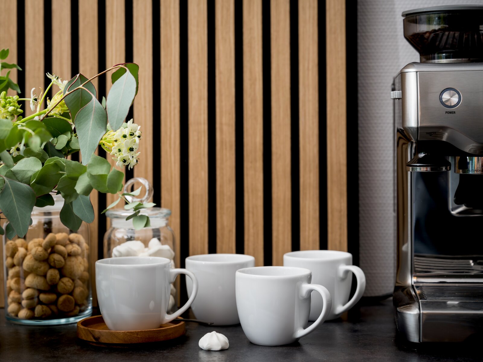 Drei weiße Kaffeetassen auf einer dunklen Küchenarbeitsplatte neben einer Edelstahl-Kaffeemaschine, mit Glasbehältern und grünem Blumenarrangement vor Holzlamellenwand, Aufnahme aus frontaler Perspektive auf Augenhöhe.