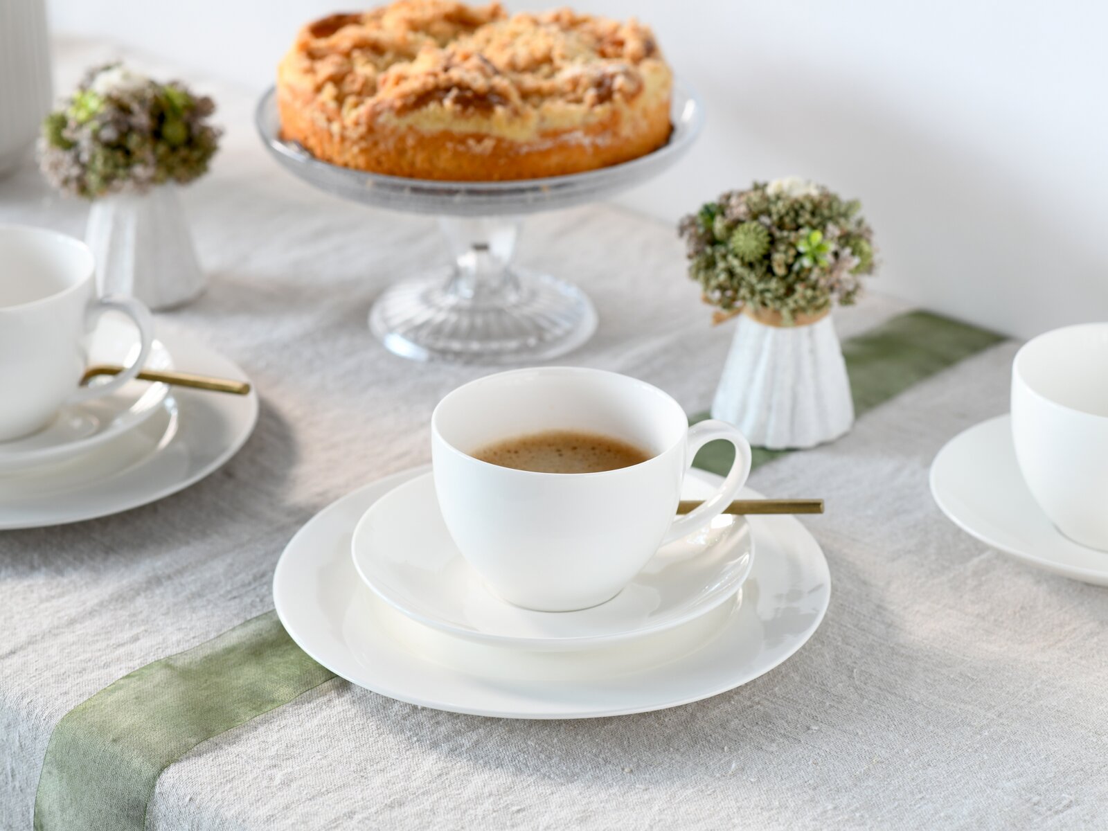 Weiße Kaffeetasse mit Untertasse und Löffel auf gedecktem Tisch, im Hintergrund ein Streuselkuchen auf Glasständer; Perspektive: leicht von oben in frontaler Ansicht