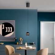 Schwarze, kugelförmige Pendelleuchte mit warmem Licht über einem Esstisch in modernem blauem Interieur, frontal aus leicht erhöhter Perspektive aufgenommen