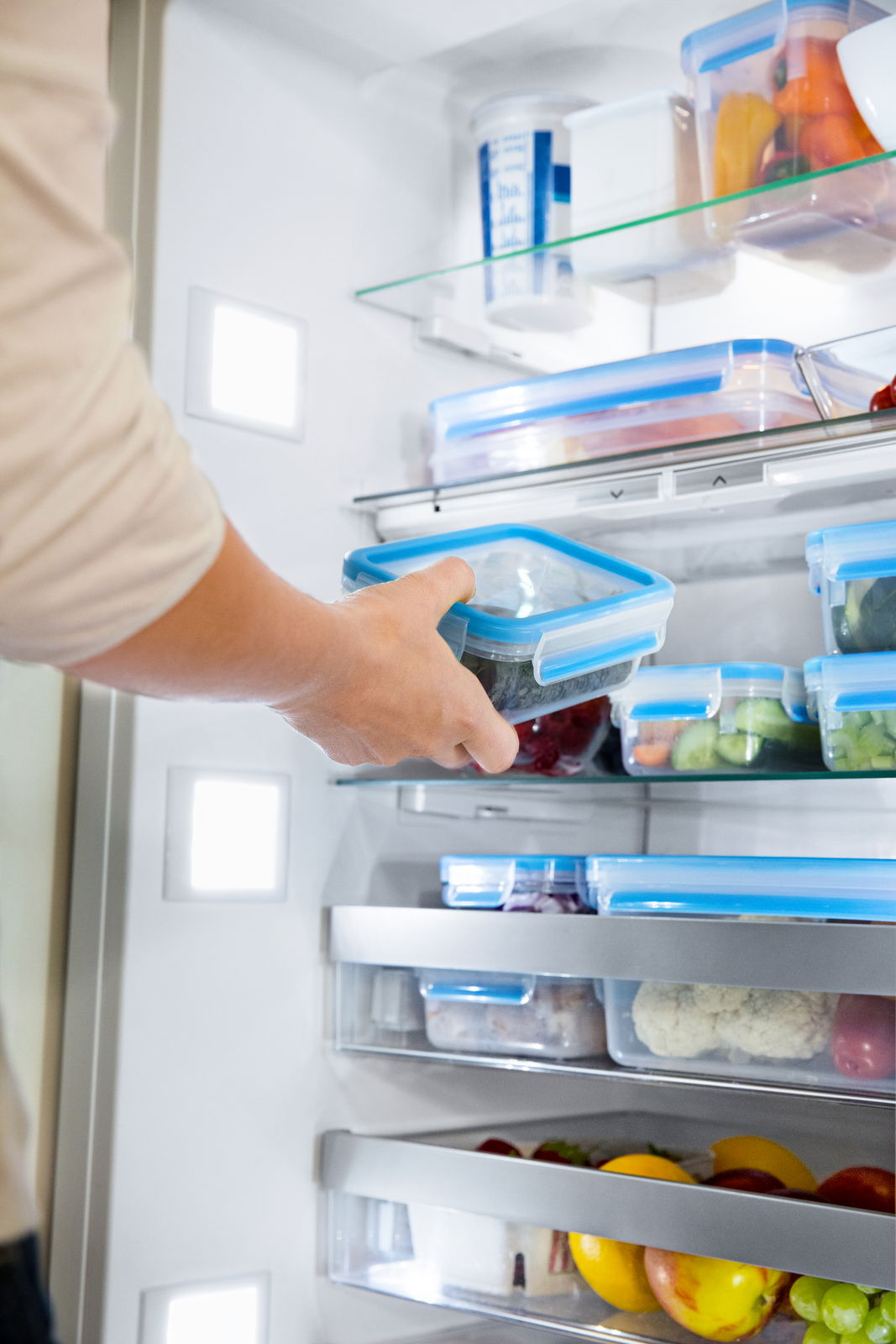 Person stellt eine rechteckige Frischhaltedose mit blauem Deckel in einen Kühlschrank, seitliche Perspektive.