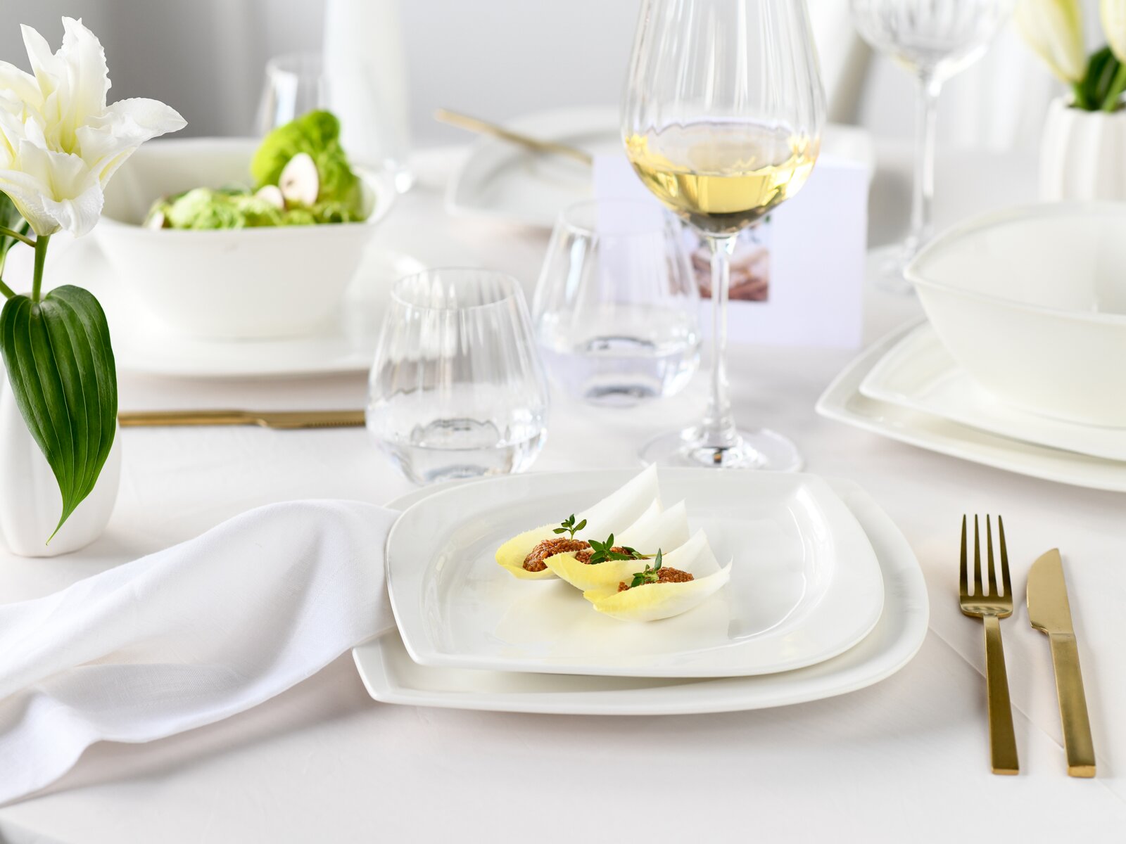 Elegant gedeckter Restauranttisch mit weißem Porzellan, Vorspeise auf quadratischem Teller, Weinglas und Wassergläsern, goldfarbenem Besteck und weißer Blume, aus leicht erhöhter seitlicher Perspektive