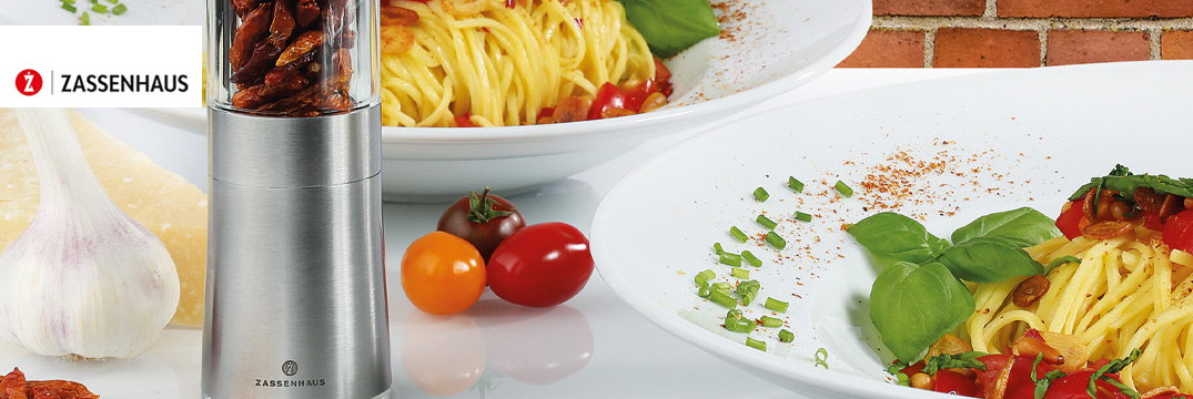 Twee borden met spaghetti, tomaten en basilicum naast een kruidenmolen, knoflook en Parmezaanse kaas.