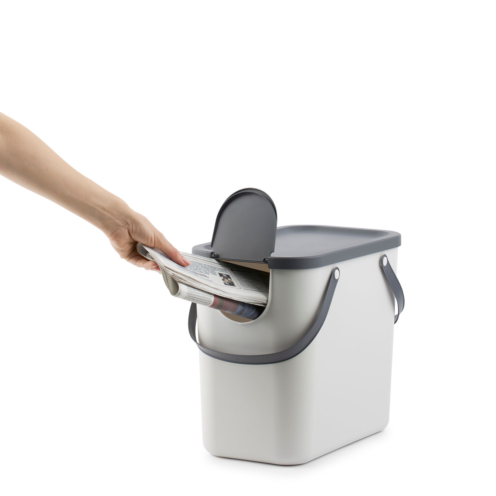 Recycling-Müllsystem mit grauem Deckel und Griffen, in das eine Hand von links eine Zeitung einwirft, seitliche Perspektive.