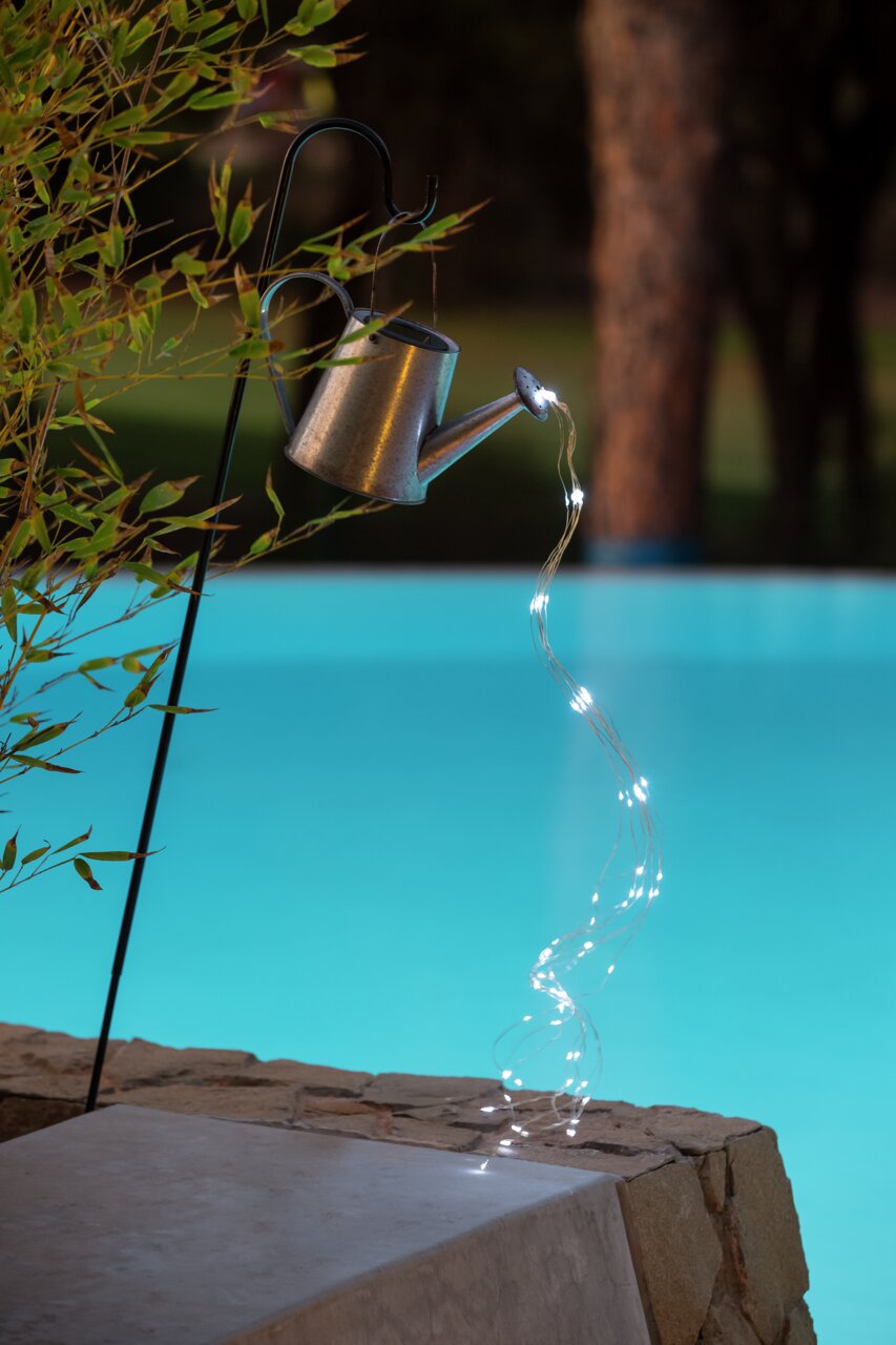 Solarleuchte in Form einer Gießkanne, die an einem Haken hängt und Lichterketten wie Wasserstrahlen ausgießt, seitliche Perspektive