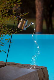 Solarleuchte in Form einer Gießkanne, die an einem Haken hängt und Lichterketten wie Wasserstrahlen ausgießt, seitliche Perspektive