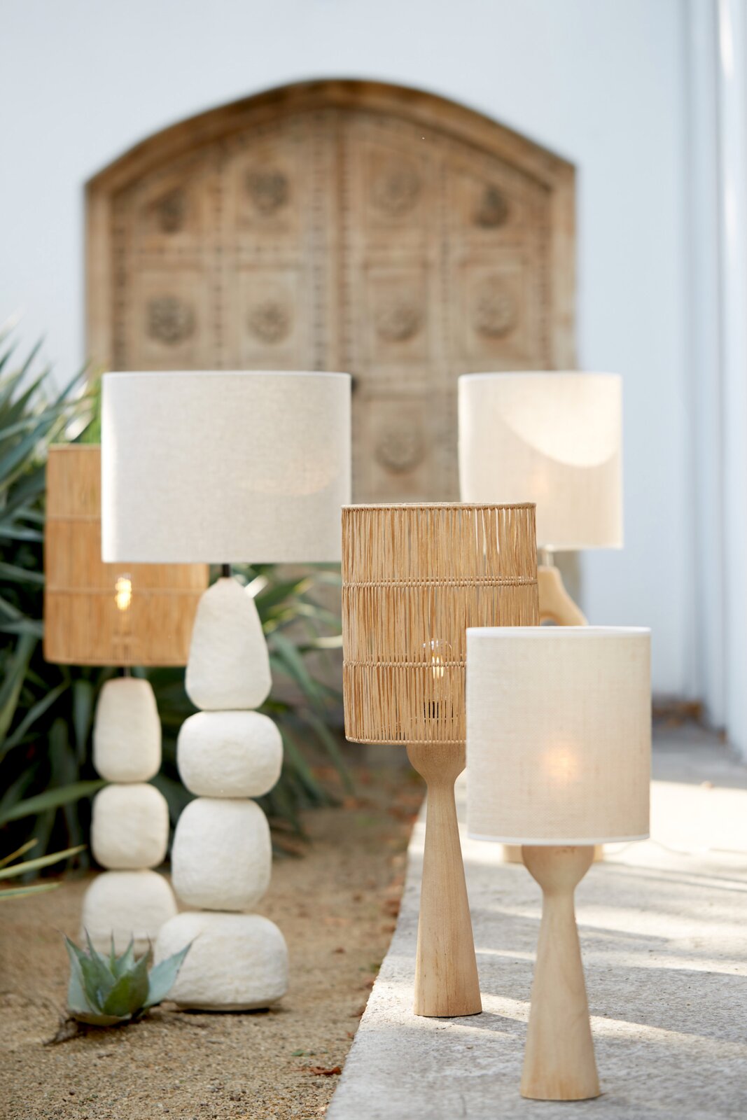 Verschillende staande lampen van natuurlijke materialen, waaronder hout en rotan, in een buitenruimte met planten en een houten deur op de achtergrond. Perspectief vanuit een schuine hoek aan de voorkant.