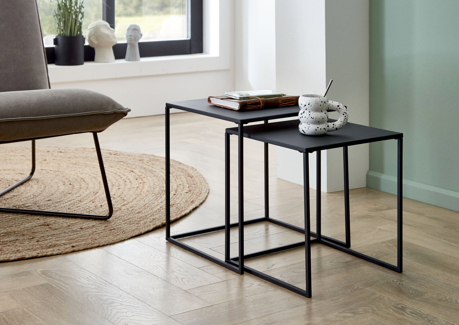 Zwei ineinandergeschobene, minimalistische Beistelltische aus schwarzem Metall im Wohnzimmer, seitliche Perspektive; auf der oberen Platte liegen ein Buch und ein Tablett, auf der vorderen Platte steht eine gesprenkelte Tasse mit Löffel.