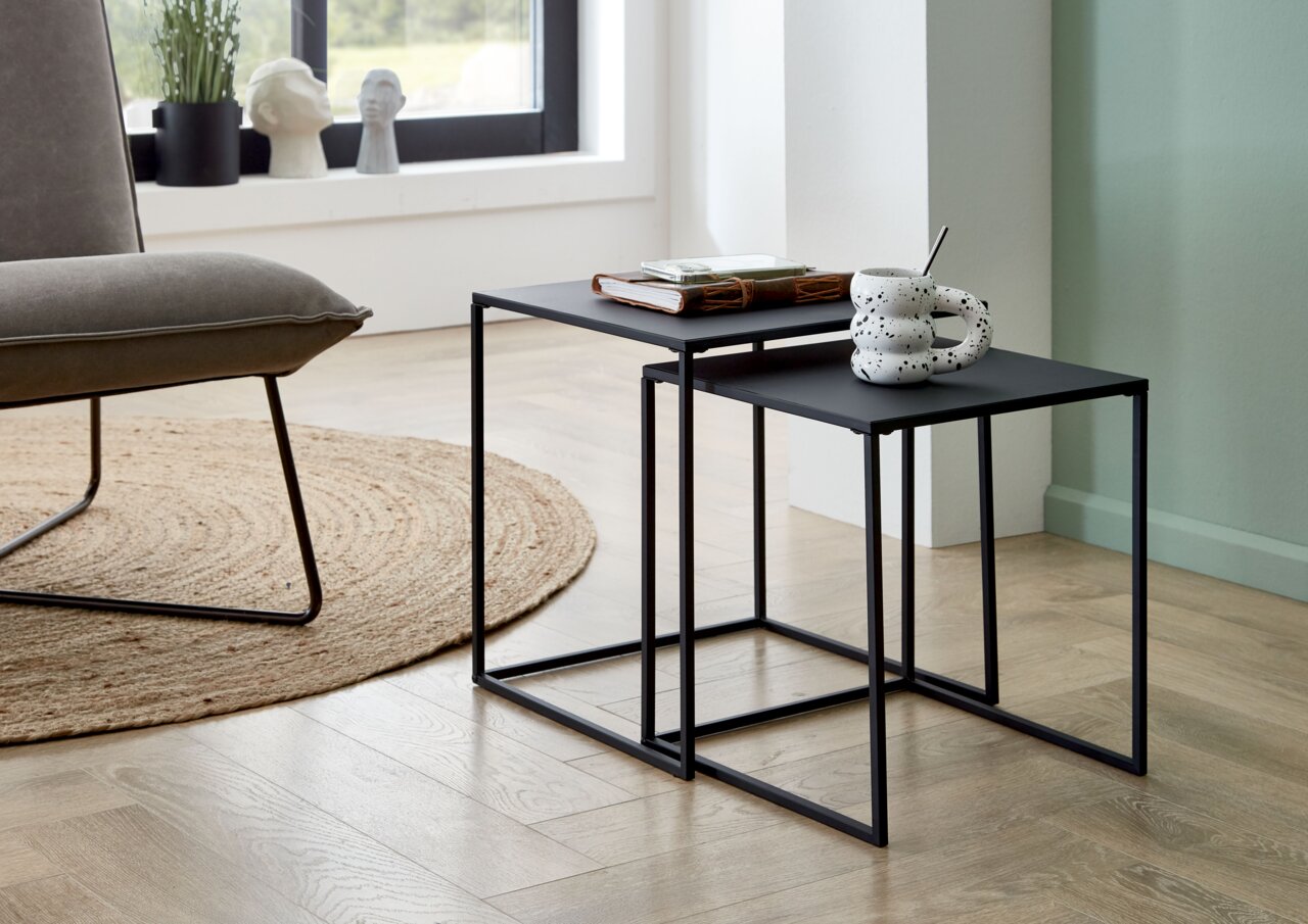 Zwei ineinandergeschobene, minimalistische Beistelltische aus schwarzem Metall im Wohnzimmer, seitliche Perspektive auf der oberen Platte liegen ein Buch und ein Tablett, auf der vorderen Platte steht eine gesprenkelte Tasse mit Löffel.