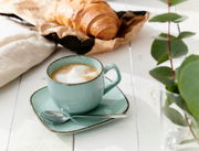 Espressotasse Casa 80ml in Blau mit Untertasse und Löffel, gefüllt mit Cappuccino, auf einem weißen Holztisch, im Hintergrund ein Croissant und grüne Blätter, seitliche Perspektive