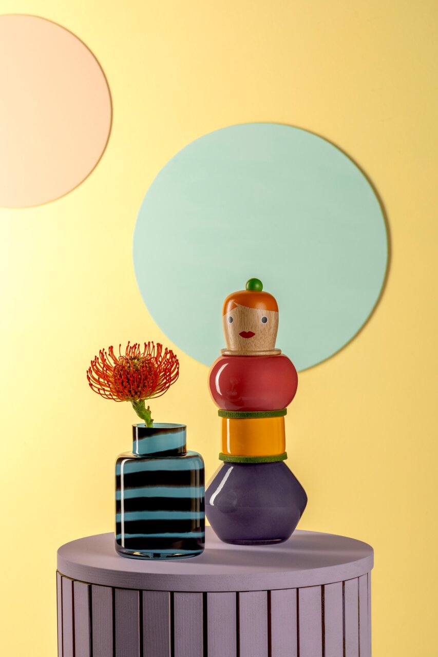 Leonardo Objektvase Simonetta BUONI AMICI Bunte Objektvase Simonetta auf einem lila Tisch, mit einer gestreiften Vase und einer Blume, vor einem gelben Hintergrund mit Kreisen.