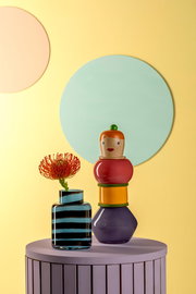 Bunte Objektvase Simonetta auf einem lila Tisch, mit einer gestreiften Vase und einer Blume, vor einem gelben Hintergrund mit Kreisen.