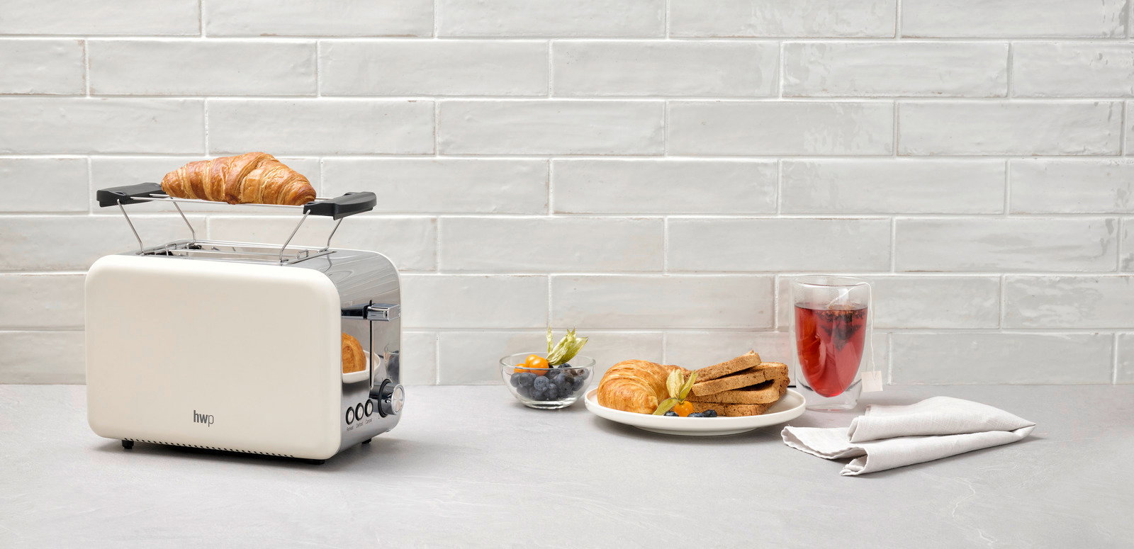 Toaster in Seitenansicht mit Croissant auf dem Brötchenaufsatz, daneben ein Teller mit Toast und Beeren sowie ein Glas Tee.
