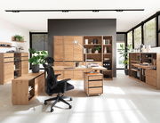 Moderne Büromöbel in einem hellen Raum mit großen Fenstern, bestehend aus einem Schreibtisch, Rollcontainer, Regalen und einem ergonomischen Bürostuhl. Perspektive von vorne.