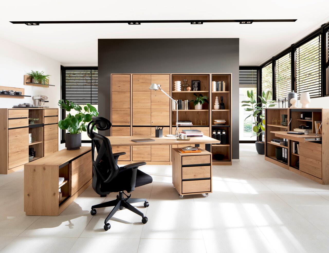 Moderne Büromöbel in einem hellen Raum mit großen Fenstern, bestehend aus einem Schreibtisch, Rollcontainer, Regalen und einem ergonomischen Bürostuhl. Perspektive von vorne.