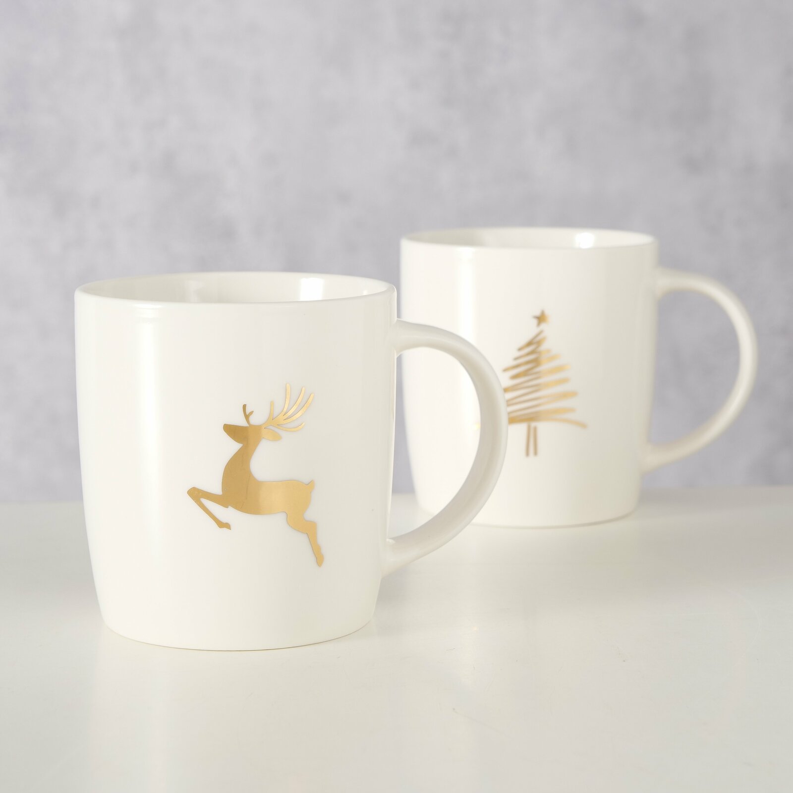Zwei weiße Becher mit goldenen Motiven, ein Rentier und ein Weihnachtsbaum, auf einer hellen Oberfläche. Vorderansicht.