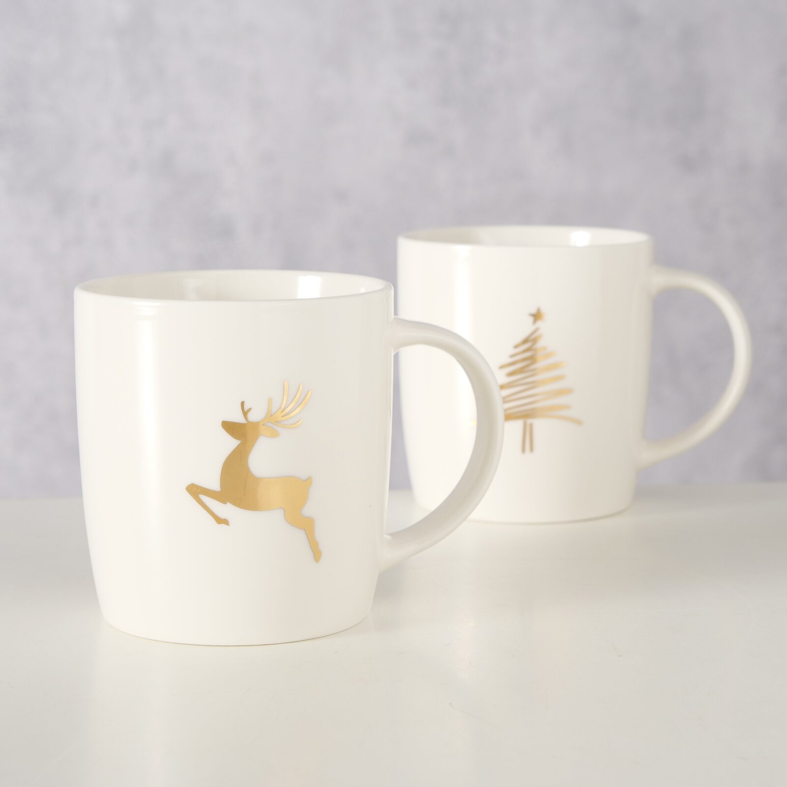 Twee witte bekers met gouden motieven, een rendier en een kerstboom, op een lichte ondergrond. Vooraanzicht.