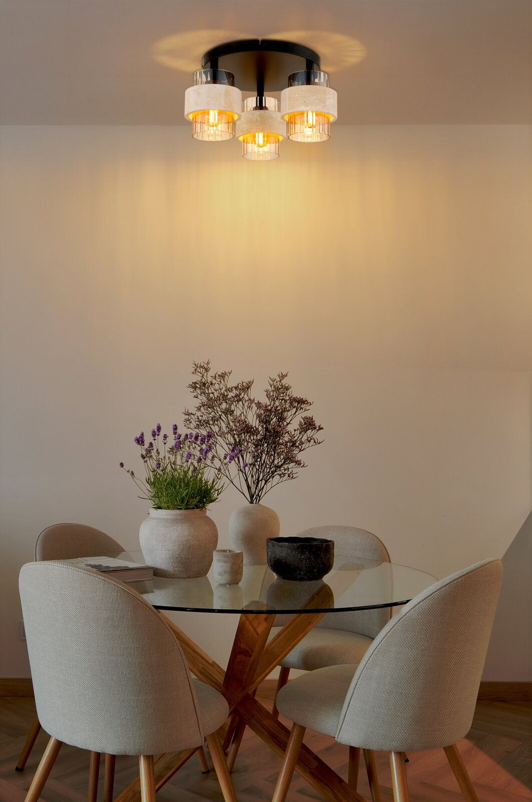 Moderne eetkamer met een rond glazen tafelblad en een houten onderstel, vier gestoffeerde stoelen en decoratieve vazen; daarboven een ingeschakelde plafondlamp met drie glazen kappen, frontaal gefotografeerd vanuit een iets lager perspectief.