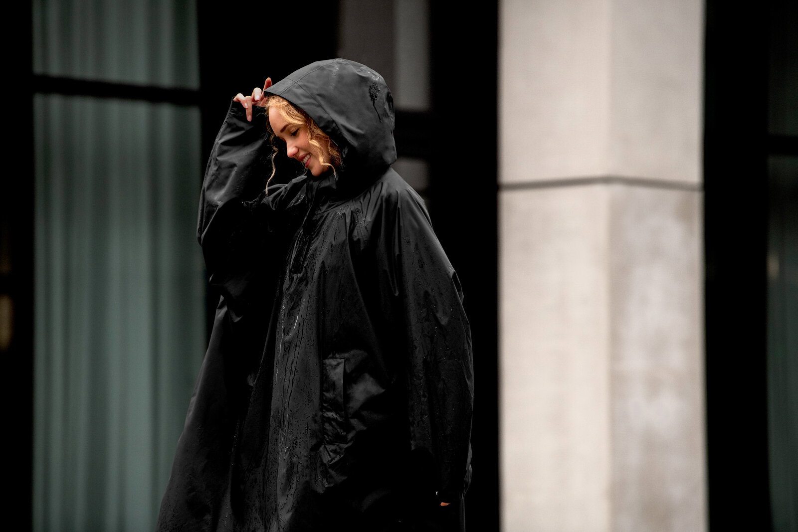 Schwarzer, wasserdichter Regenponcho mit Kapuze, von der Seite in leichter Untersicht fotografiert, getragen von einer Person im Regen vor einem modernen Gebäude.