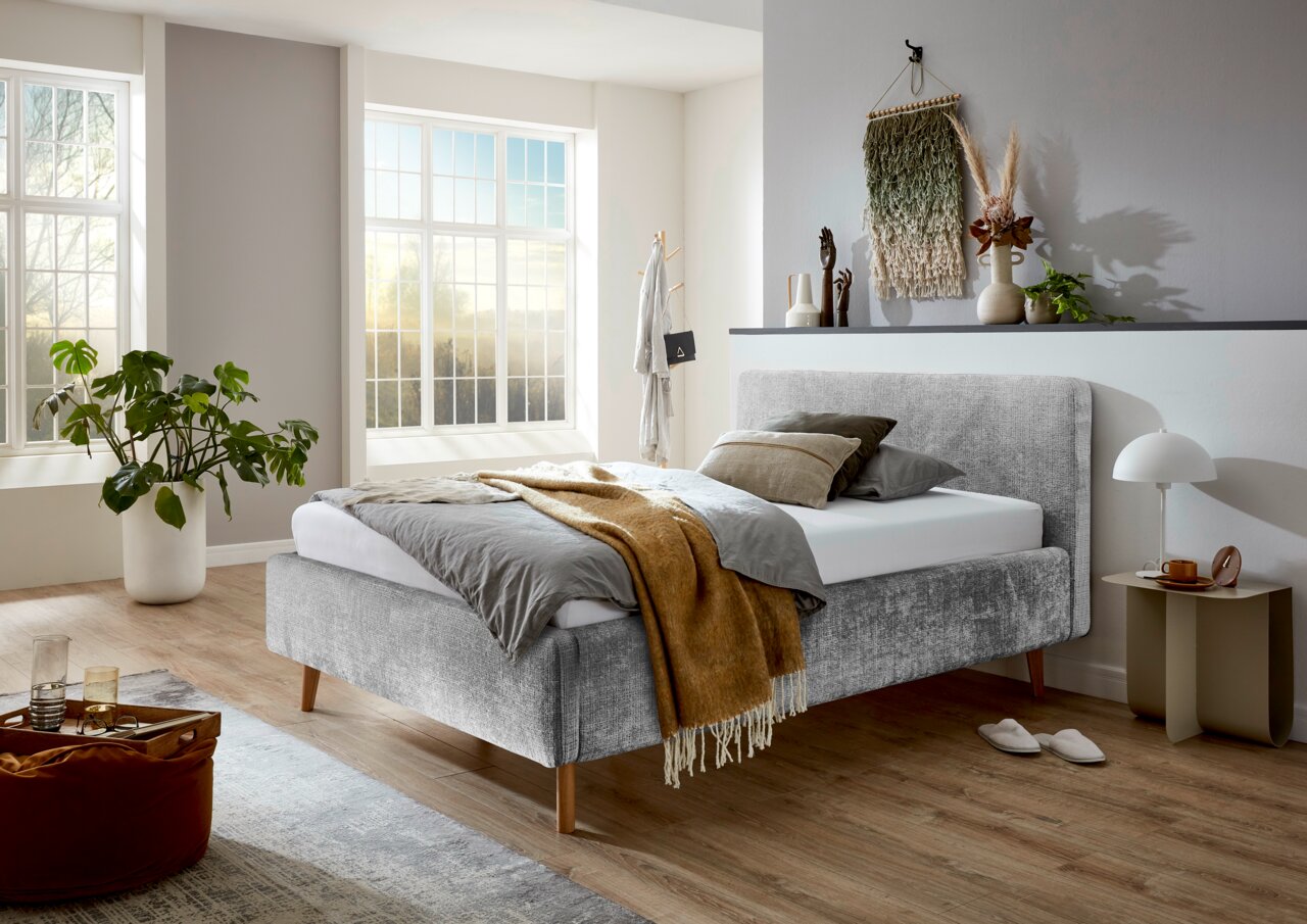 Modern gestoffeerd bedframe in een lichte slaapkamer met grote ramen, zijaanzicht.