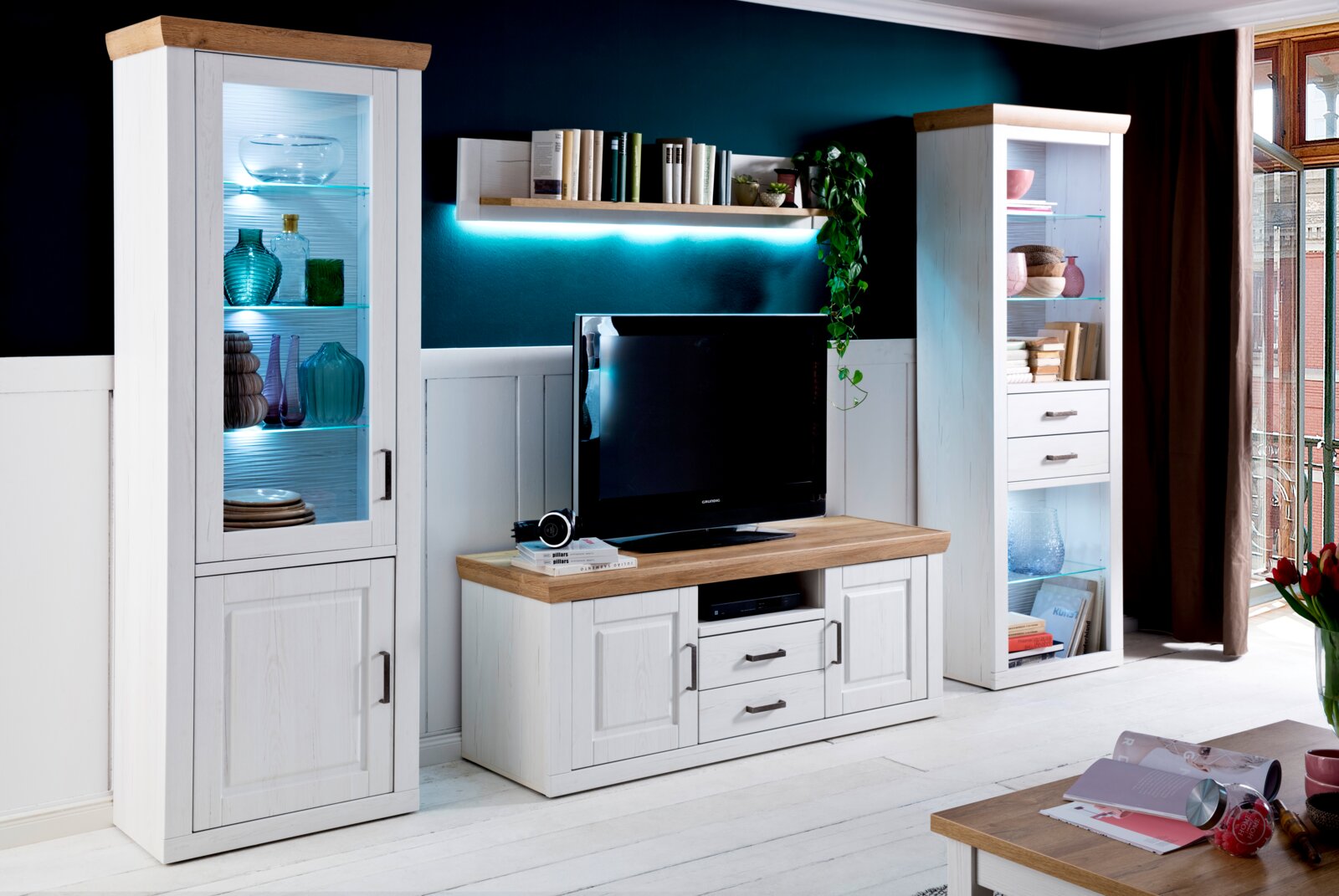 Landscape Wandkombination BREGENZ Wohnzimmermöbel-Set im Landhausstil mit weißer Holzoptik und Eichenholz-Absetzungen: links beleuchtete Glasvitrine, mittig TV-Lowboard und Wandboard mit LED-Licht, rechts hohe Vitrine mit Schubladen; Frontansicht aus leicht schräger Perspektive.