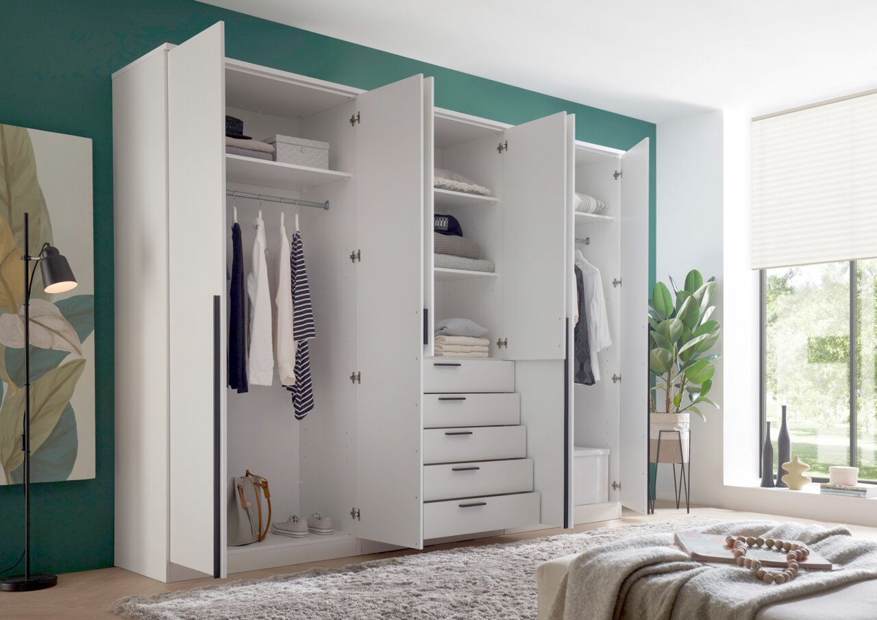 Moderner, weißer Drehtürenschrank mit sechs Türen, teilweise geöffnet, zeigt Innenfächer, Kleiderstangen und Schubladen. Perspektive von vorne links.