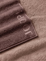 Nahaufnahme aus schräger Draufsicht: zwei flauschige Handtücher in Taupe- und Beigetönen, das obere mit eingewebtem JOOP!-Logo und eingefasster Kante.