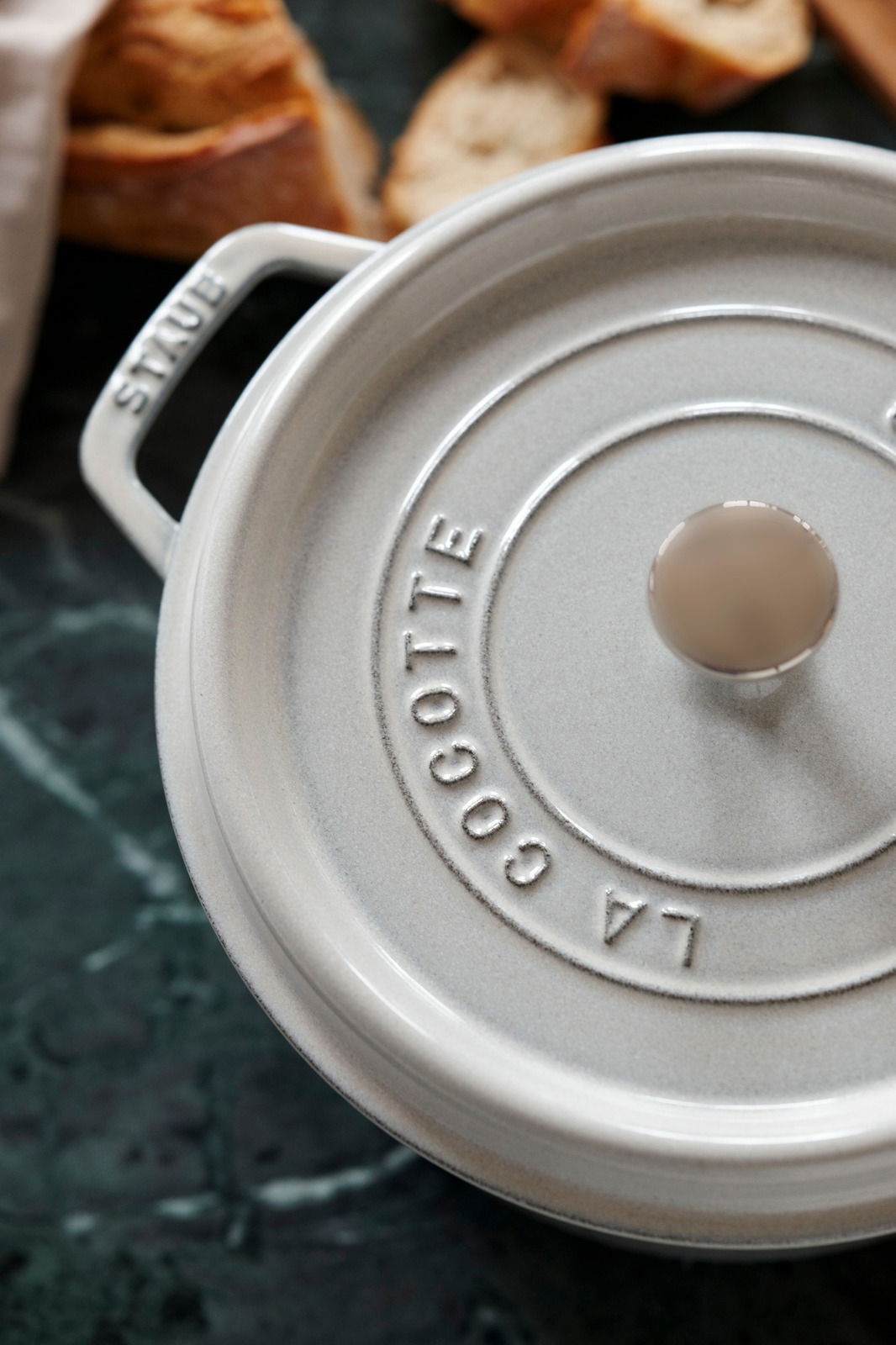 Obere Perspektive auf einen runden, grauen Bräter Cocotte mit Deckel und Griffen, umgeben von Brotstücken.