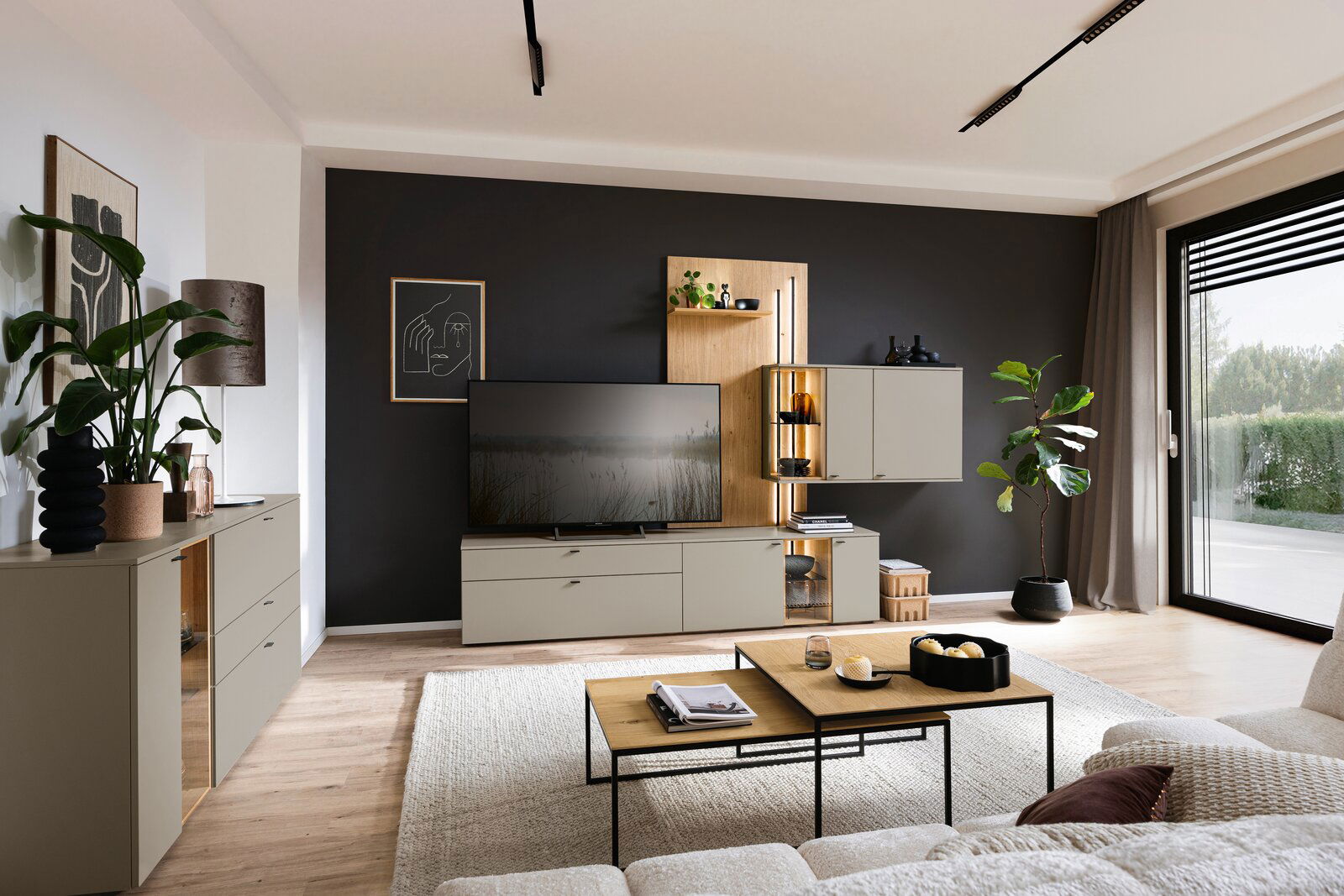 Moderne Wohnzimmer-Wandkombination Venjakob VANYA in einem stilvollen Raum mit schwarzer Akzentwand, TV, Pflanzen und dekorativen Elementen, seitliche Perspektive.
