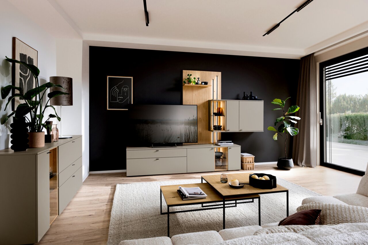 Moderne Wohnzimmer-Wandkombination Venjakob VANYA in einem stilvollen Raum mit schwarzer Akzentwand, TV, Pflanzen und dekorativen Elementen, seitliche Perspektive.
