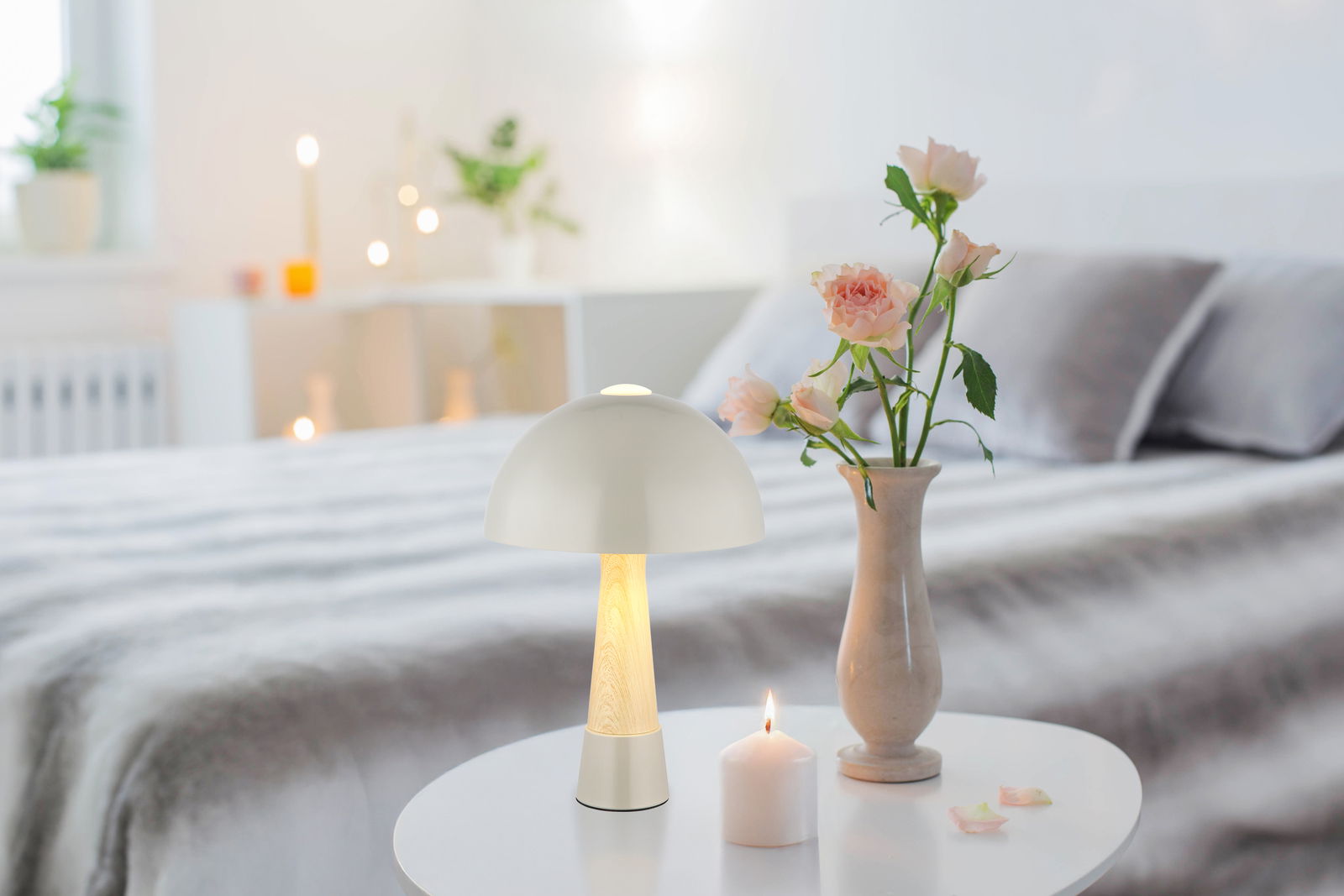 Moderne Tischleuchte mit weißem Schirm und Holzfuß auf einem Tisch, umgeben von einer Vase mit Rosen und einer brennenden Kerze, seitliche Perspektive.