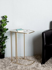 Moderner Beistelltisch mit goldfarbenem Metallgestell und transparenter Glasplatte neben schwarzem Sofa auf Teppich, Frontansicht leicht von oben.
