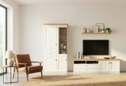Frontale Ansicht eines hellen Wohnzimmers mit weißer Wohnwand im Landhausstil: hoher Vitrinenschrank mit Holzplatte und TV-Lowboard mit Holzoberfläche unter einem Wandfernseher.