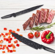 Zwei schwarze Messer mit gesprenkelter Klinge auf einem weißen Holztisch, umgeben von geschnittenem Steak, Paprikawürfeln, Tomaten und einem Rosmarinzweig. Draufsicht.