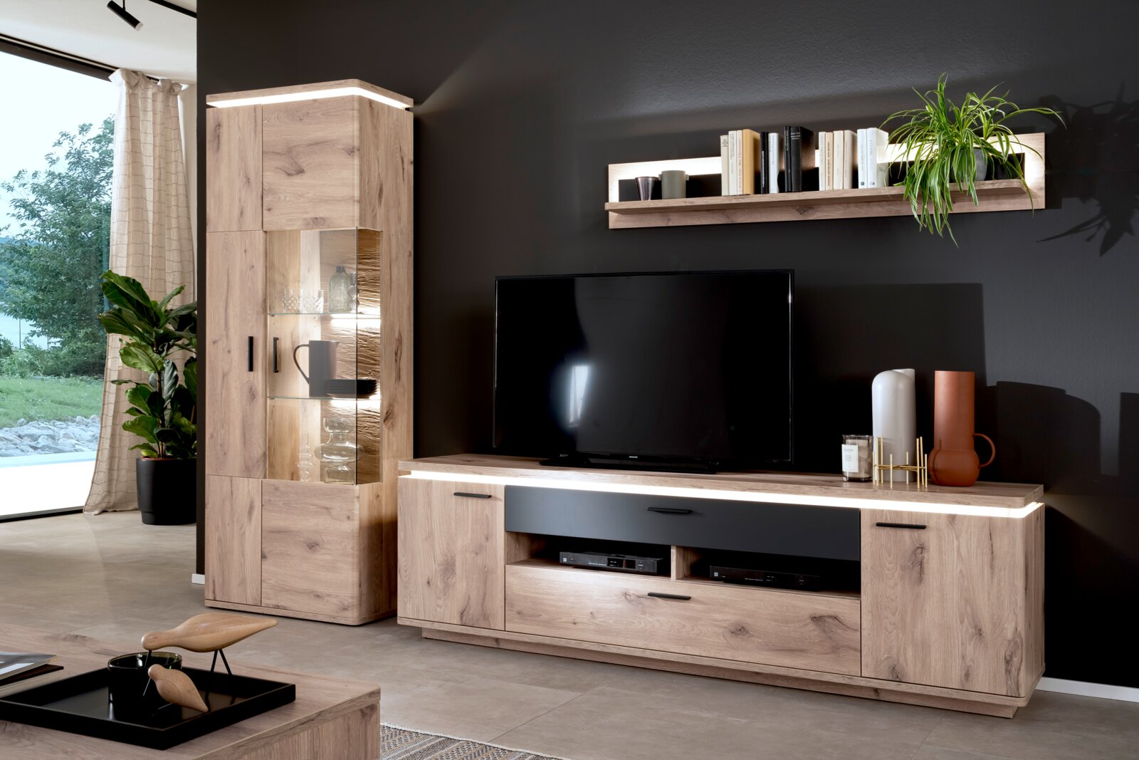 Zurbrüggen Wandcombinatie MODENA Woonkamerwand in eikenlook met tv-lowboard, vitrinekast met glazen deuren en wandplank, led-verlichting en zwarte accenten, gefotografeerd vanuit een vooraanzicht, lichtjes van links.