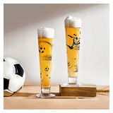 Zwei hohe Biergläser mit Fußballmotiven, gefüllt mit Bier und Schaumkrone, auf einer Holzunterlage und neben einem Fußball, seitliche Perspektive.