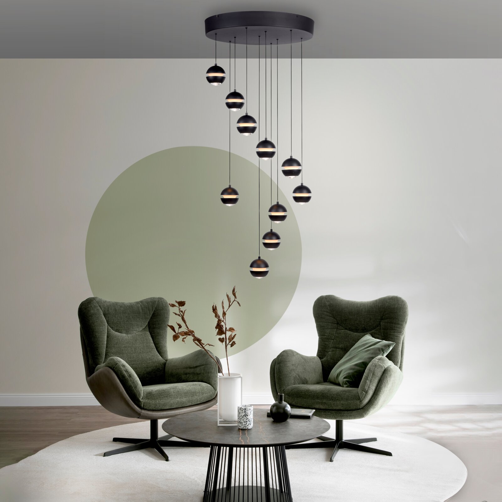 Moderne zwarte hanglamp met meerdere bolvormige lampenkappen aan een ronde plafondplaat, van voren gezien licht van onderaf in de woonkamer, met op de achtergrond twee groene fauteuils.