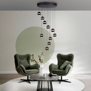 Moderne zwarte hanglamp met meerdere bolvormige lampenkappen aan een ronde plafondplaat, van voren gezien licht van onderaf in de woonkamer, met op de achtergrond twee groene fauteuils.