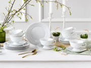 Weißes Porzellan-Geschirrset mit Tassen, Untertassen und Tellern im klassischen Reliefdesign, dekoriert mit Frühlingszweigen und Kerzenleuchtern, fotografiert in frontaler Tischansicht leicht von oben.
