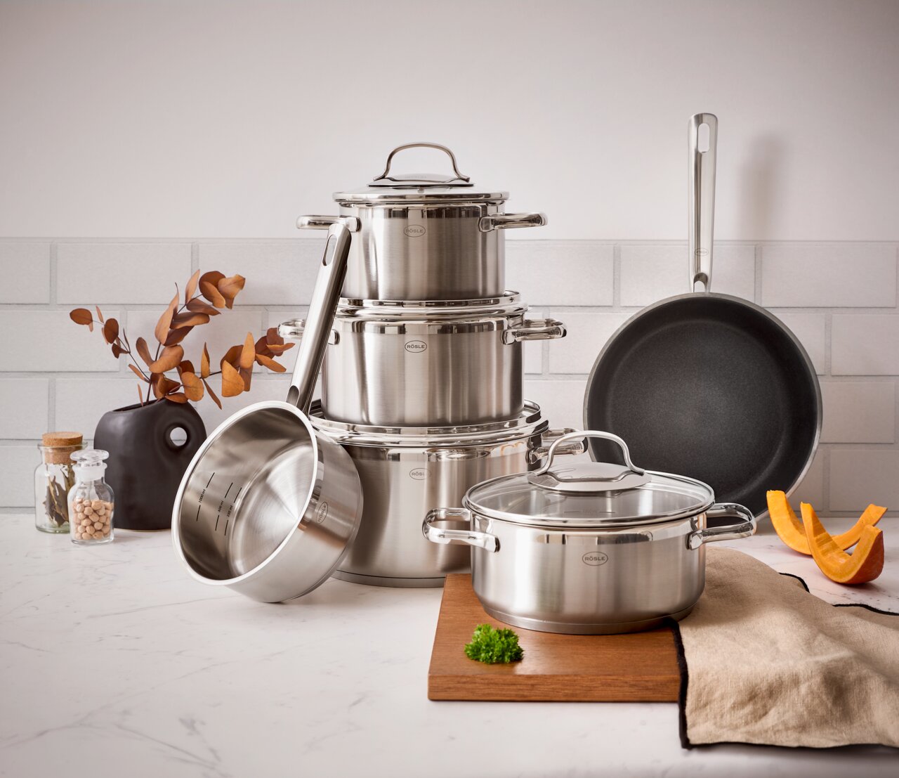 Topfset ELEGANCE 6-tlg. aus Edelstahl, bestehend aus mehreren Töpfen und einer Pfanne, auf einer Küchenarbeitsplatte arrangiert. Perspektive von vorne.